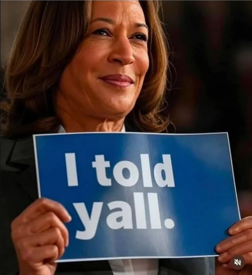 A woman with brown hair and a suit holds a blue sign with white text that reads 'I told yall.' She has a confident, slightly…