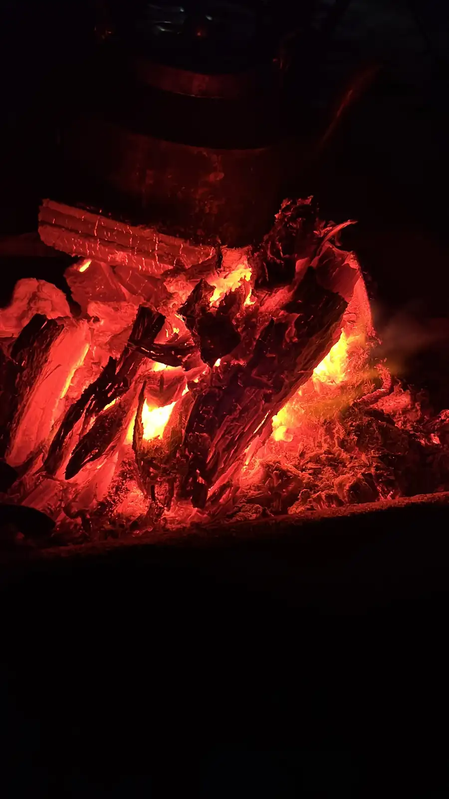 A close-up photo of glowing red and orange embers and charred wood in a dark fireplace or grill, with a large metal pot or…