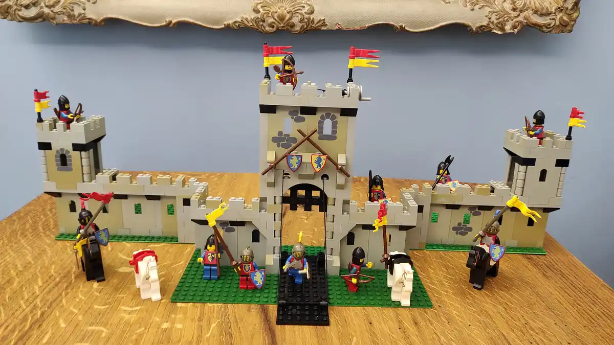 A detailed LEGO castle with multiple towers, flags, and minifigures in medieval armor, some on horseback, arranged on a…