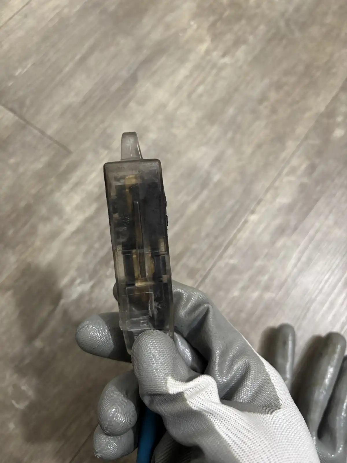 A hand wearing a gray work glove holds a transparent plastic object with internal metal parts, resembling a gun, against a…