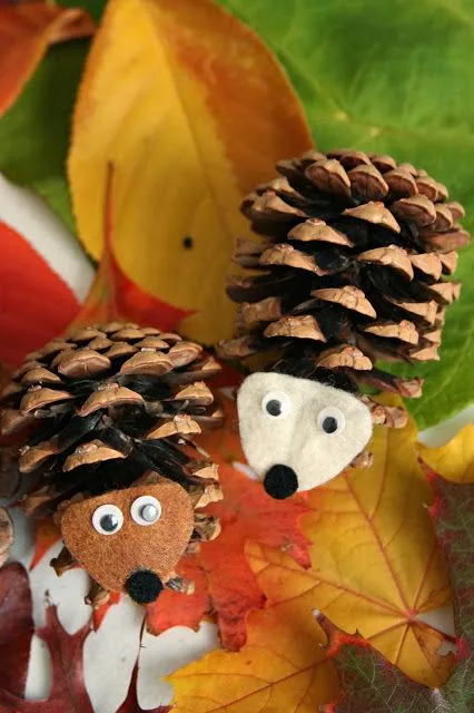 Two pinecones are decorated as hedgehogs with googly eyes, felt snouts, and black pom-pom noses, sitting among colorful…