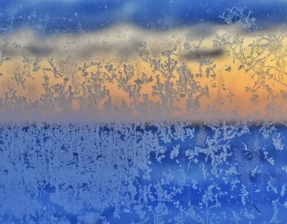 A frost-covered window shows a sunrise with orange and blue hues, while intricate ice patterns obscure the view. Overlaid…