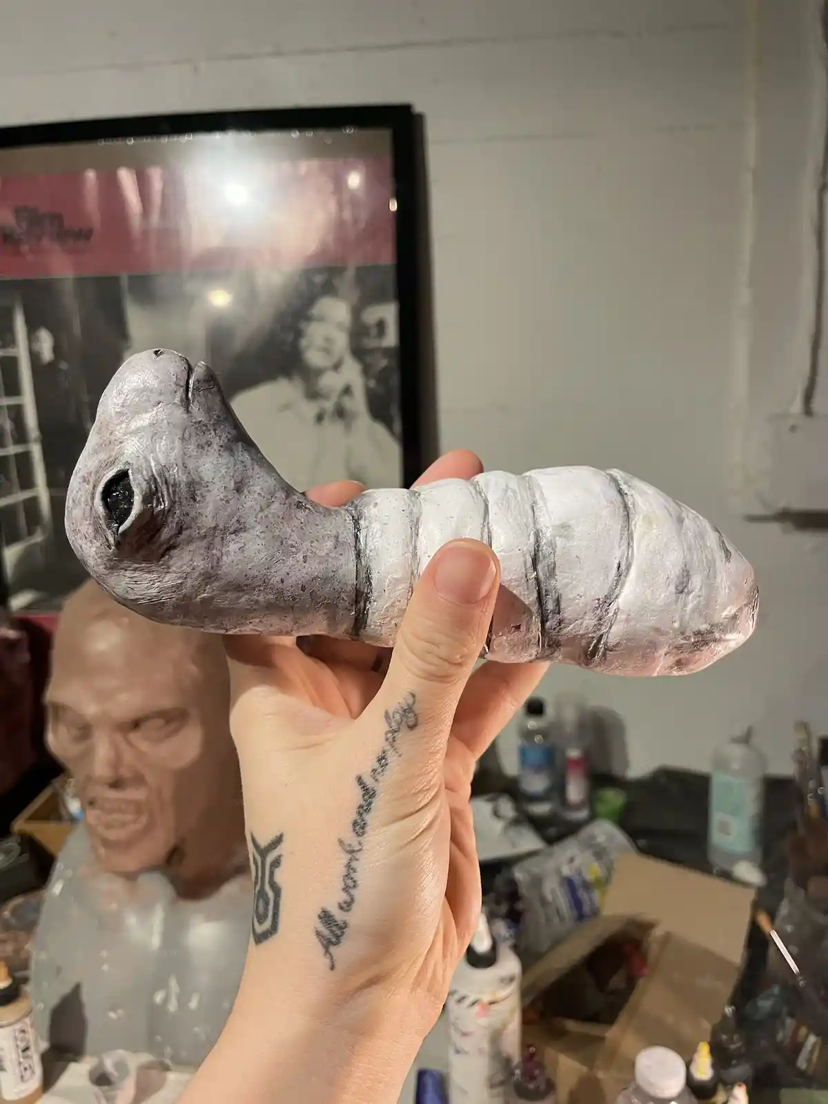 A hand holds a handmade, silver-painted alien-like creature with a large eye and segmented body. The hand has tattoos…