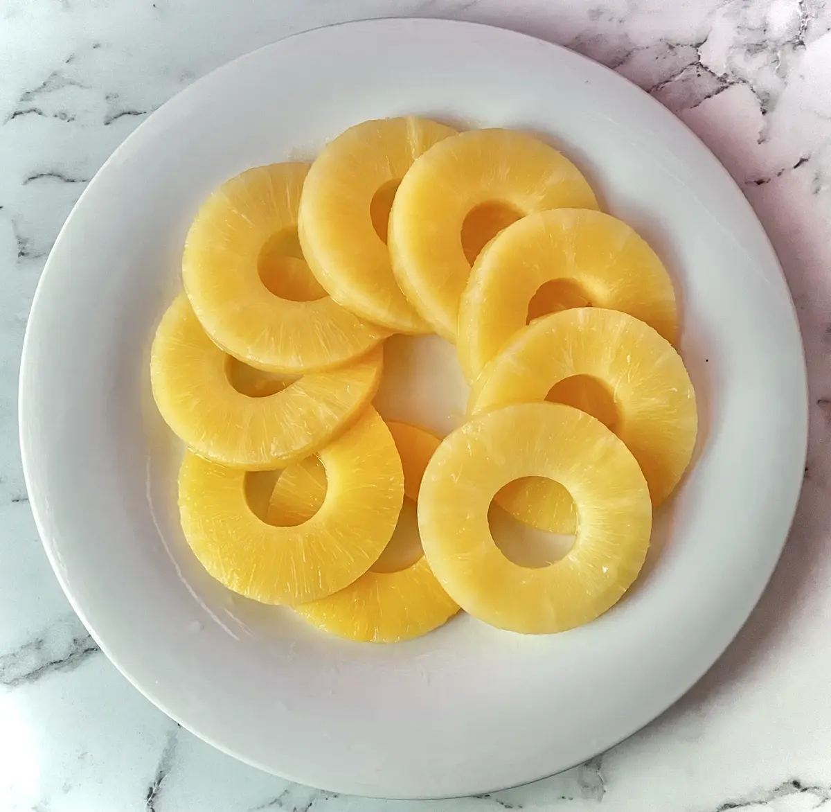 A white plate holds several bright yellow, circular pineapple rings with their centers removed, arranged on a white and gray…