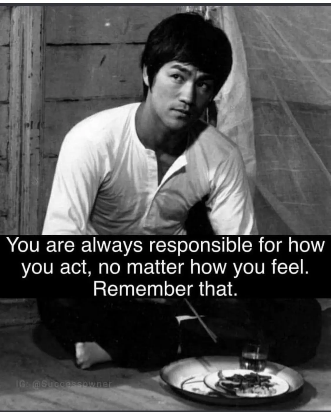 A black-and-white photo of Bruce Lee sitting cross-legged on the floor, looking intently at the viewer. In front of him is a…