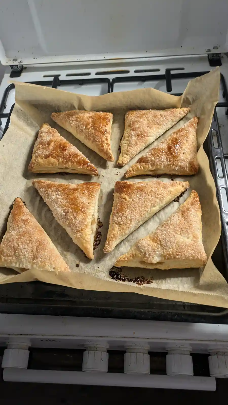 A baking tray lined with parchment paper holds eight golden-brown, triangular pastries, likely turnovers or samosas, resting…