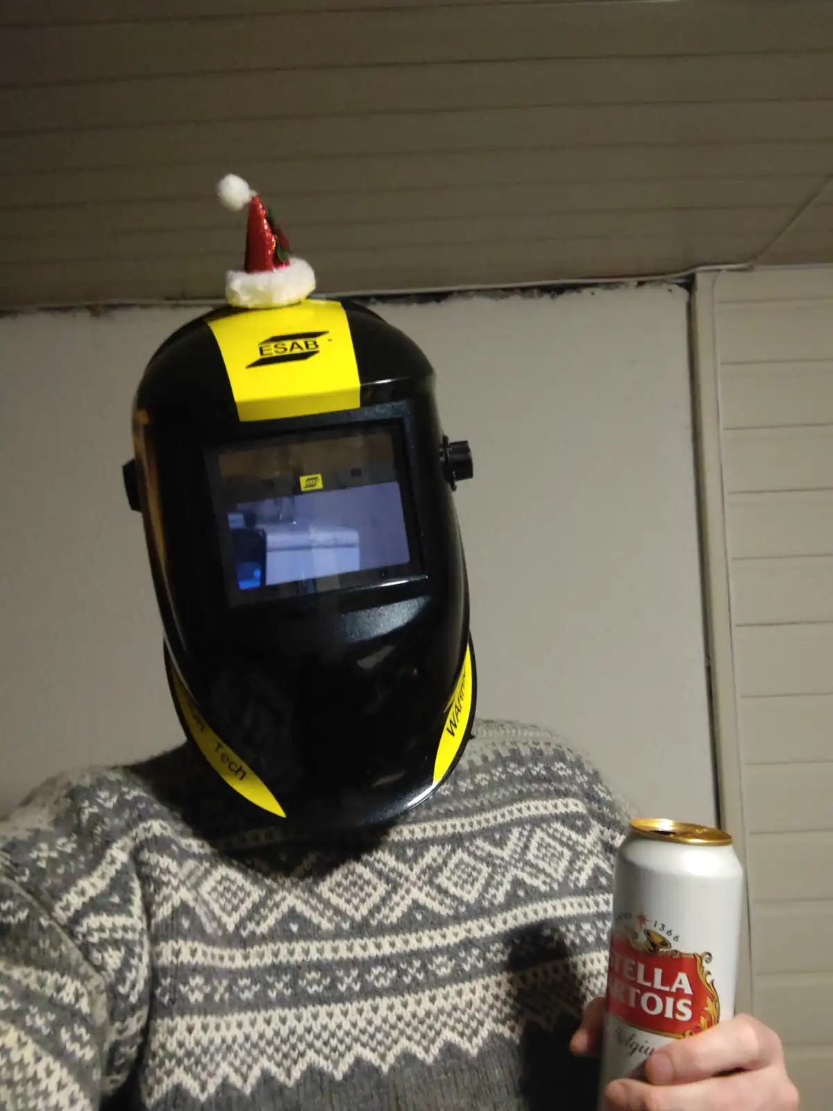 A person wearing a black and yellow ESAB welding helmet with a small red and white Santa hat perched on top, holds a can of…