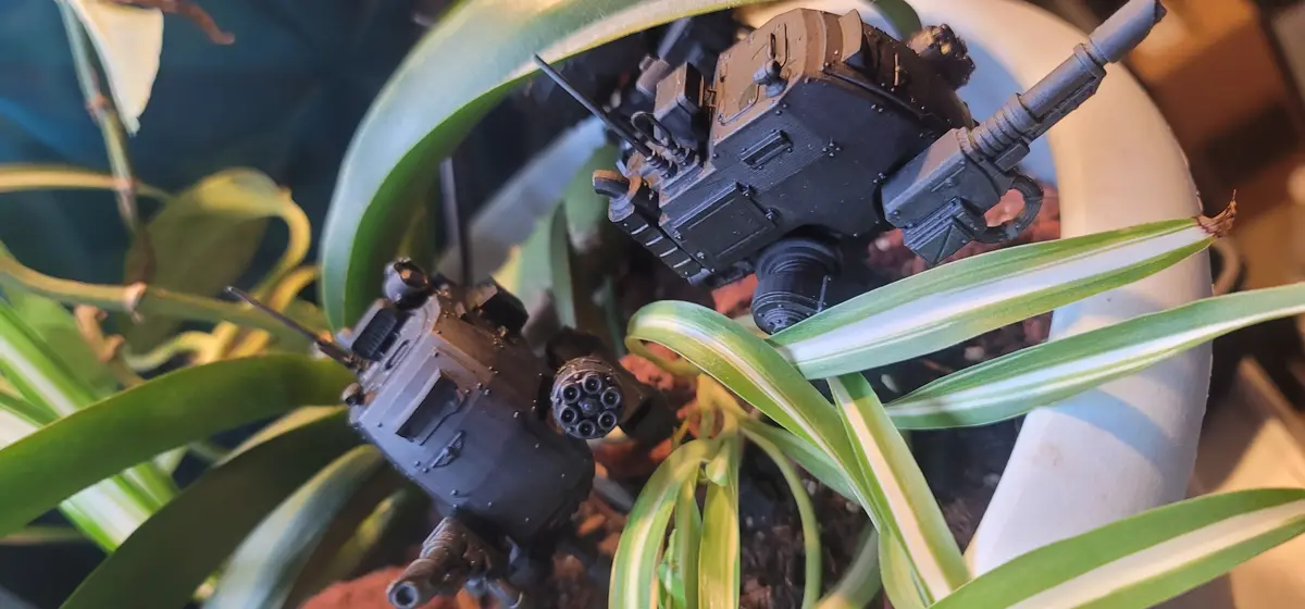 Two miniature black sci-fi tanks are nestled among the green and white striped leaves of a potted plant, appearing to be…