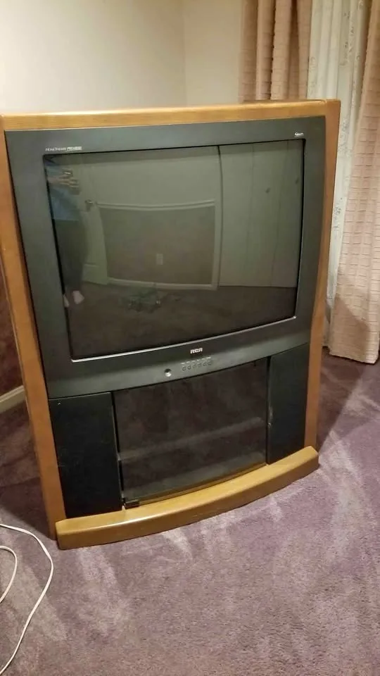 A large, old-style CRT television with a wooden frame and built-in speakers sits on a purple carpet, its screen dark and…
