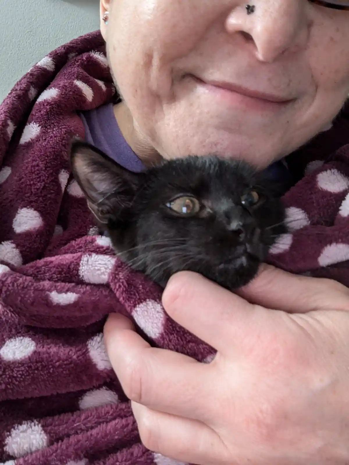 A close-up photo shows a person with a nose piercing and a maroon polka-dot blanket holding a small black kitten. The kitten…