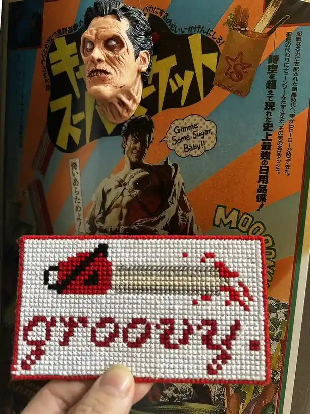 A hand holds a red-and-white cross-stitched patch depicting a pixelated chainsaw and the word 'groovy' in block letters…