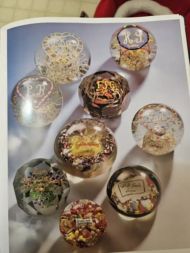 A photo of a printed page showing nine decorative glass paperweights, each with unique engravings, names, or messages in…