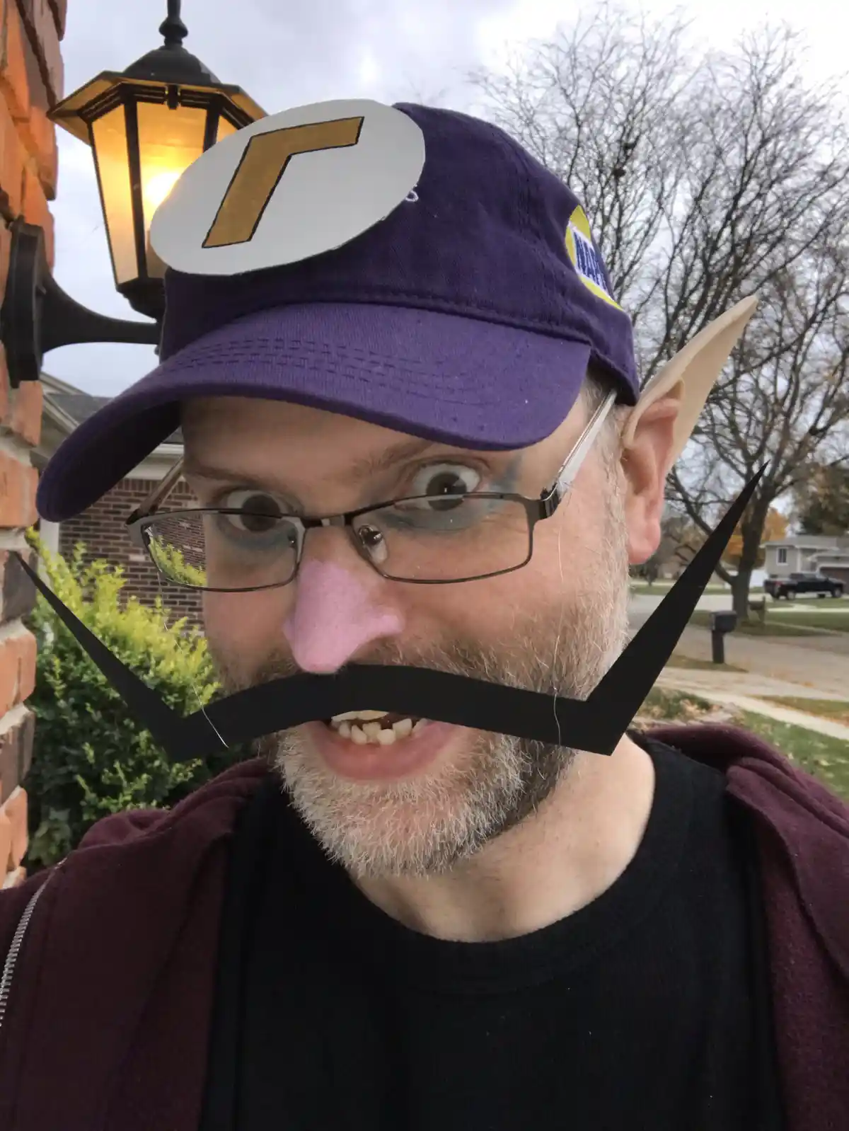 A man outdoors wears a purple cap with a white circle and gold 'R' logo, has a pink nose, black handlebar mustache, and…