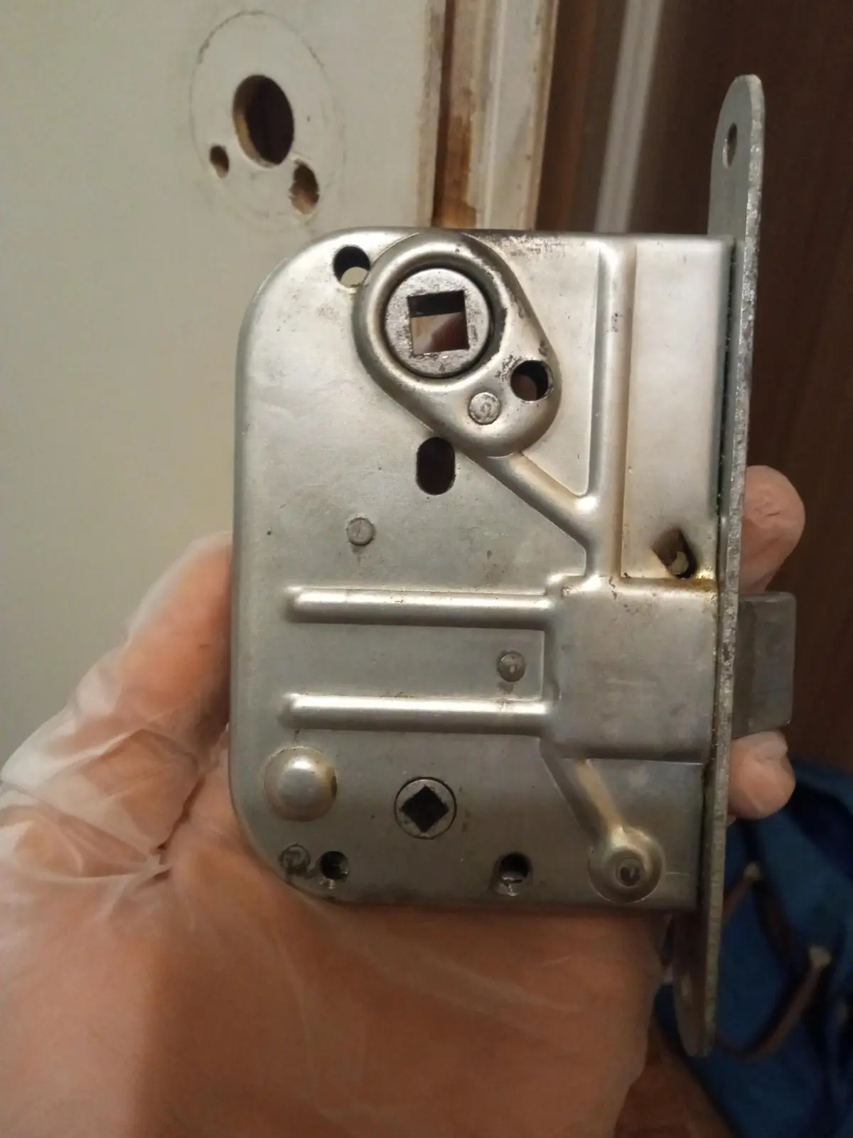 A hand wearing a clear plastic glove holds a silver metal door lock mechanism, showing its backside with screws and mounting…