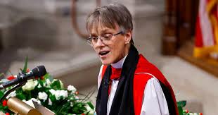 A woman in academic robes stands at a podium with a microphone, speaking with a slightly forced, polite expression. She has…