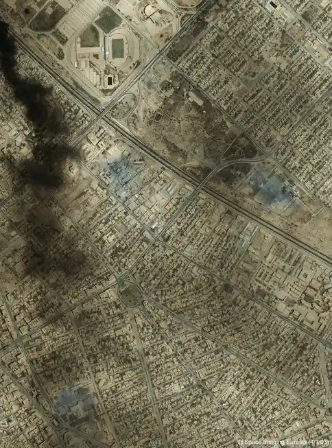 An aerial satellite image shows a sprawling urban area with grid-like streets, numerous buildings, and major roadways…