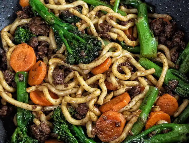 a close-up photo of a stir fry in a dark pan, featuring thick udon noodles mixed with chopped carrots, green broccoli stalks…