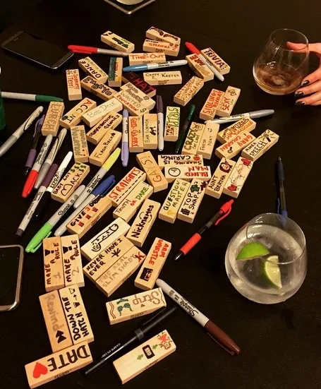 A dark table is covered with dozens of wooden dominoes, each hand-written with various names, phrases, or dates like 'DATE'…