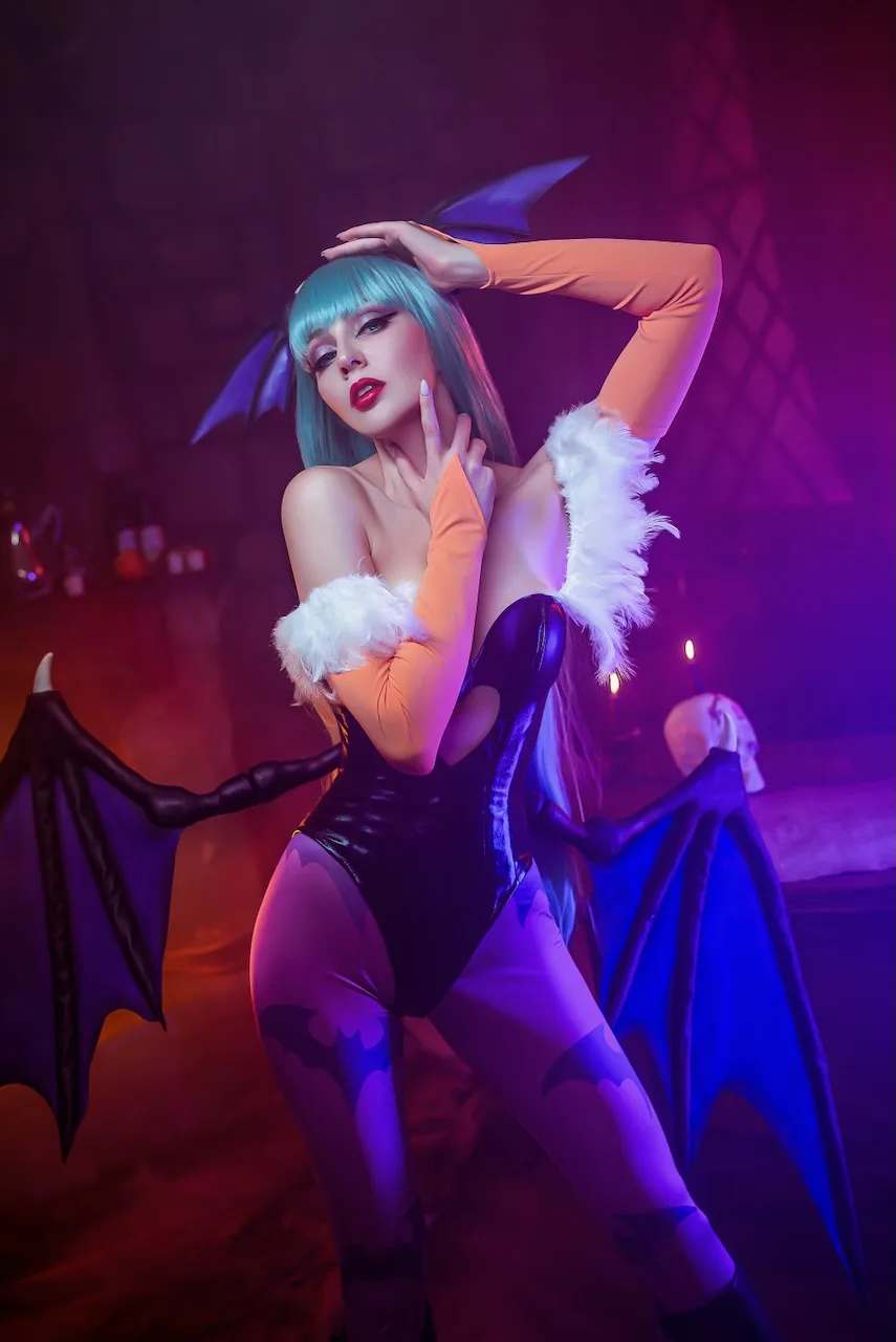 A person with blue hair and bat-like wings poses in a black bodysuit with white fur trim and bat-patterned leggings, set…
