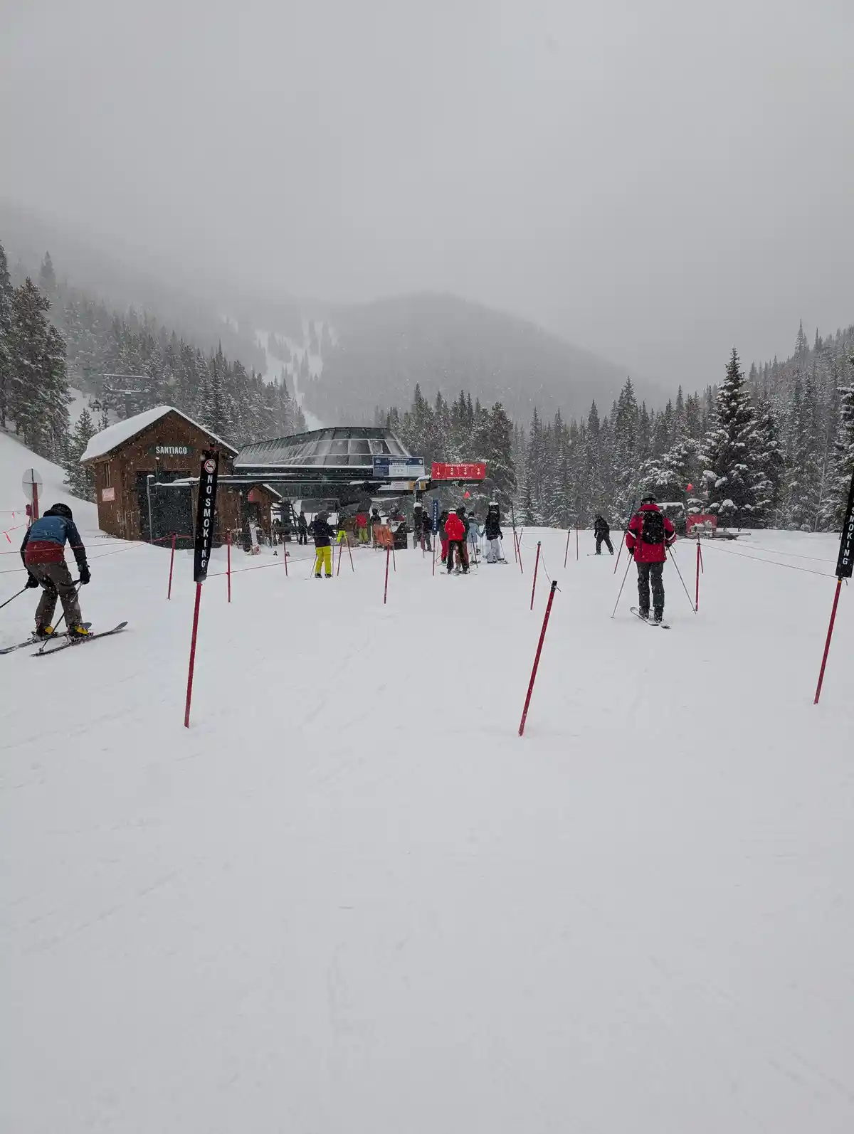 A snowy ski resort scene with skiers waiting near a lift station labeled 'SANTIAGO' under overcast skies, with red poles and…