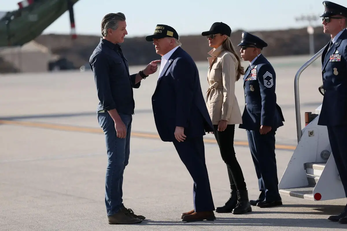 A man in a dark shirt and jeans points at Donald Trump, who wears a 'Make America Great Again' cap, while Melania Trump and…