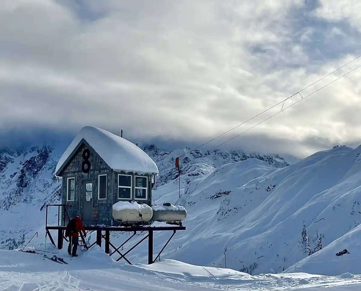 A person in a red jacket and backpack stands on snowy ground near a small wooden cabin elevated on stilts, with a large…