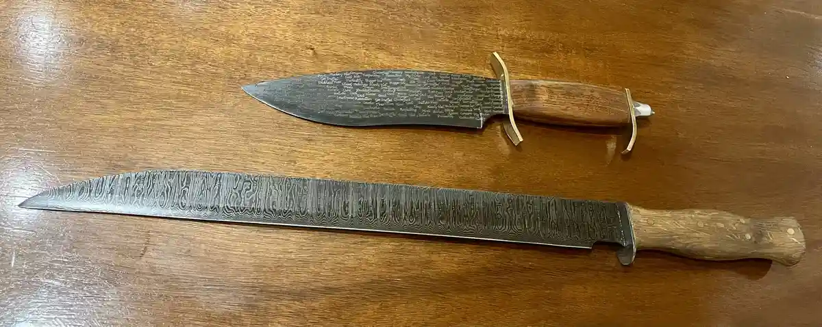 Two handmade knives with wooden handles lie on a wooden table. The top knife has a shorter blade with tiny engraved text…