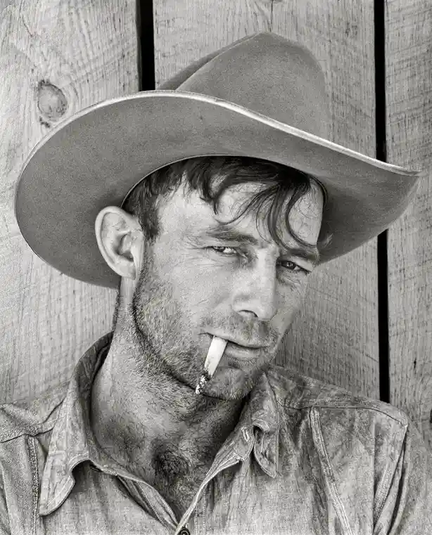 A black-and-white portrait of a man wearing a cowboy hat and denim shirt, with a cigarette in his mouth, standing against a…