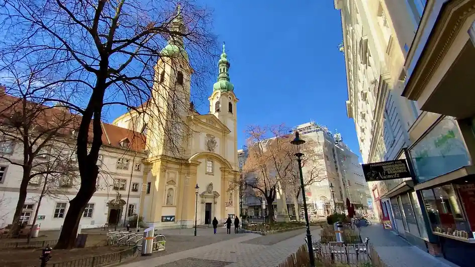 A sunny urban street scene featuring a large yellow church with green domed towers, bare trees, and pedestrians. A black…