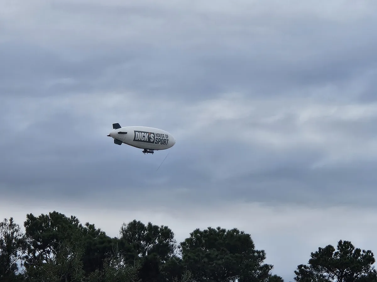 A white blimp with the text 'DICK'S HOUSE OF SPORT' is flying against a cloudy sky, with tree tops visible at the bottom of…