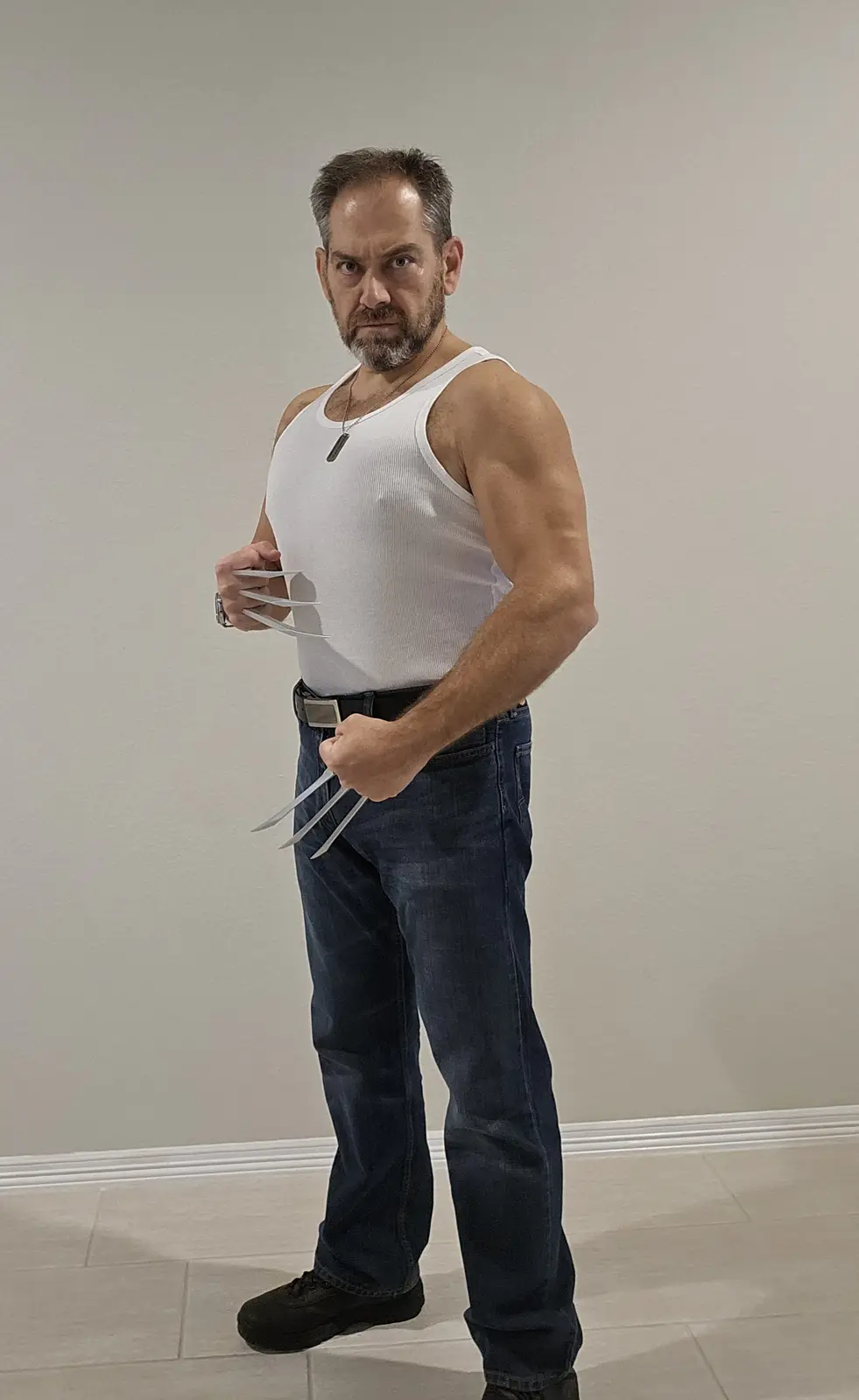 A middle-aged man with a beard and muscular build stands indoors wearing a white tank top, blue jeans, and black shoes. He…