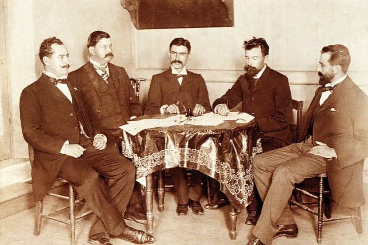 a sepia-toned historical photograph shows five men in formal early 20th-century suits seated around a table covered with a…