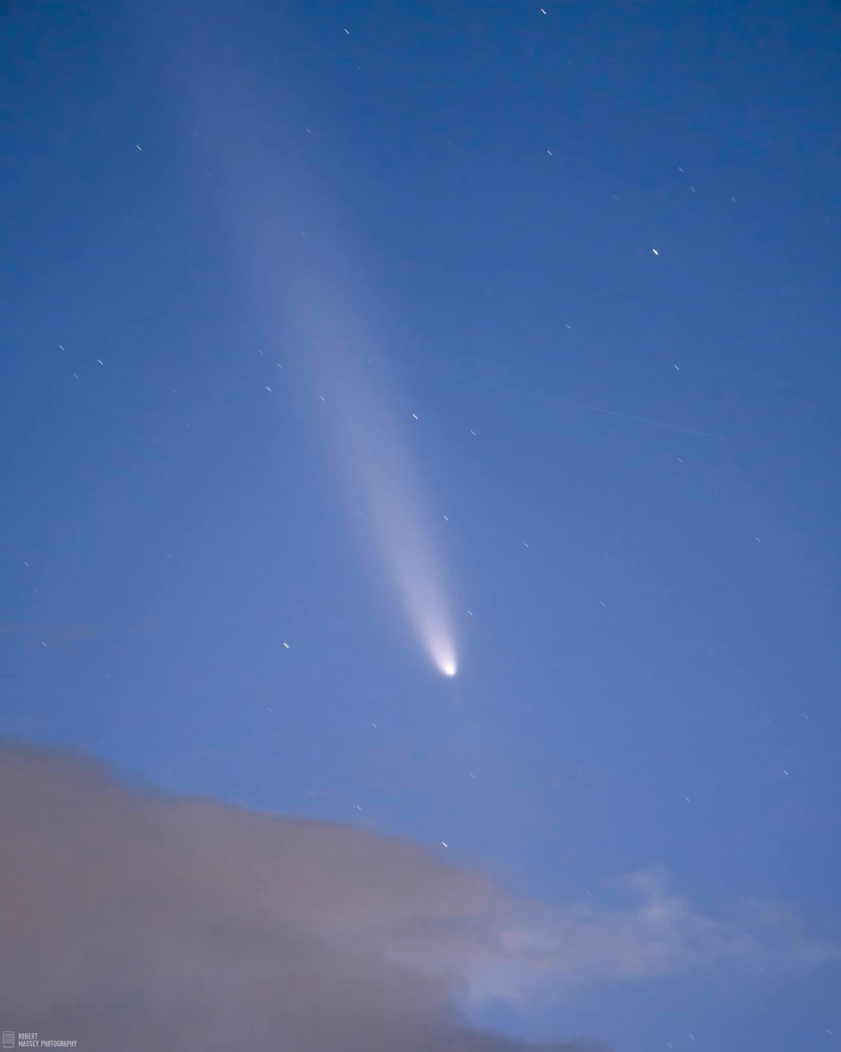 A photograph of a bright comet with a long, diffuse tail streaking across a deep blue night sky dotted with stars, with a…