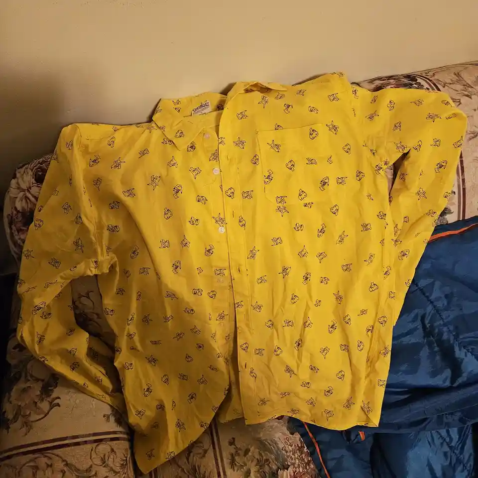 Two identical yellow button-up shirts with a repeating black outline pattern of Pikachu from Pokémon are laid out…
