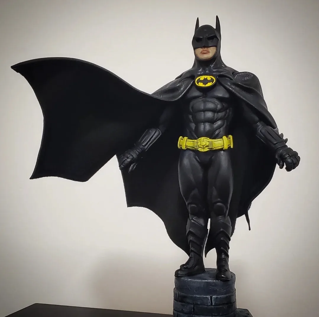 A Batman statue in a black suit with a yellow utility belt and bat symbol, standing on a stone pedestal with a flowing cape…