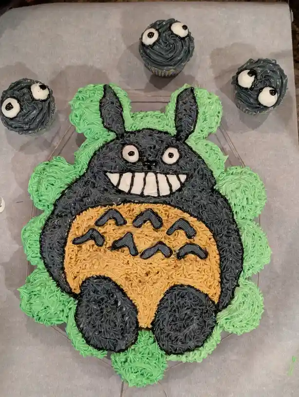 A large cake shaped and decorated like the character Totoro from My Neighbor Totoro, featuring black and tan frosting with…