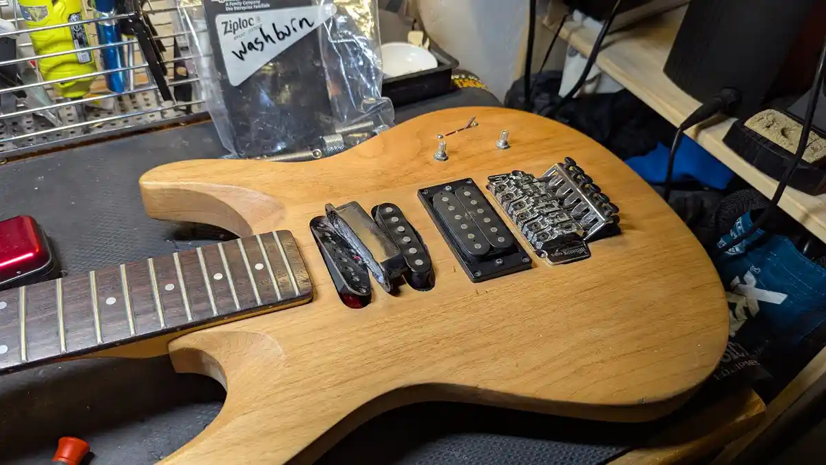 A light-wood electric guitar body sits on a workbench, its pickups and bridge removed and placed nearby, suggesting a repair…