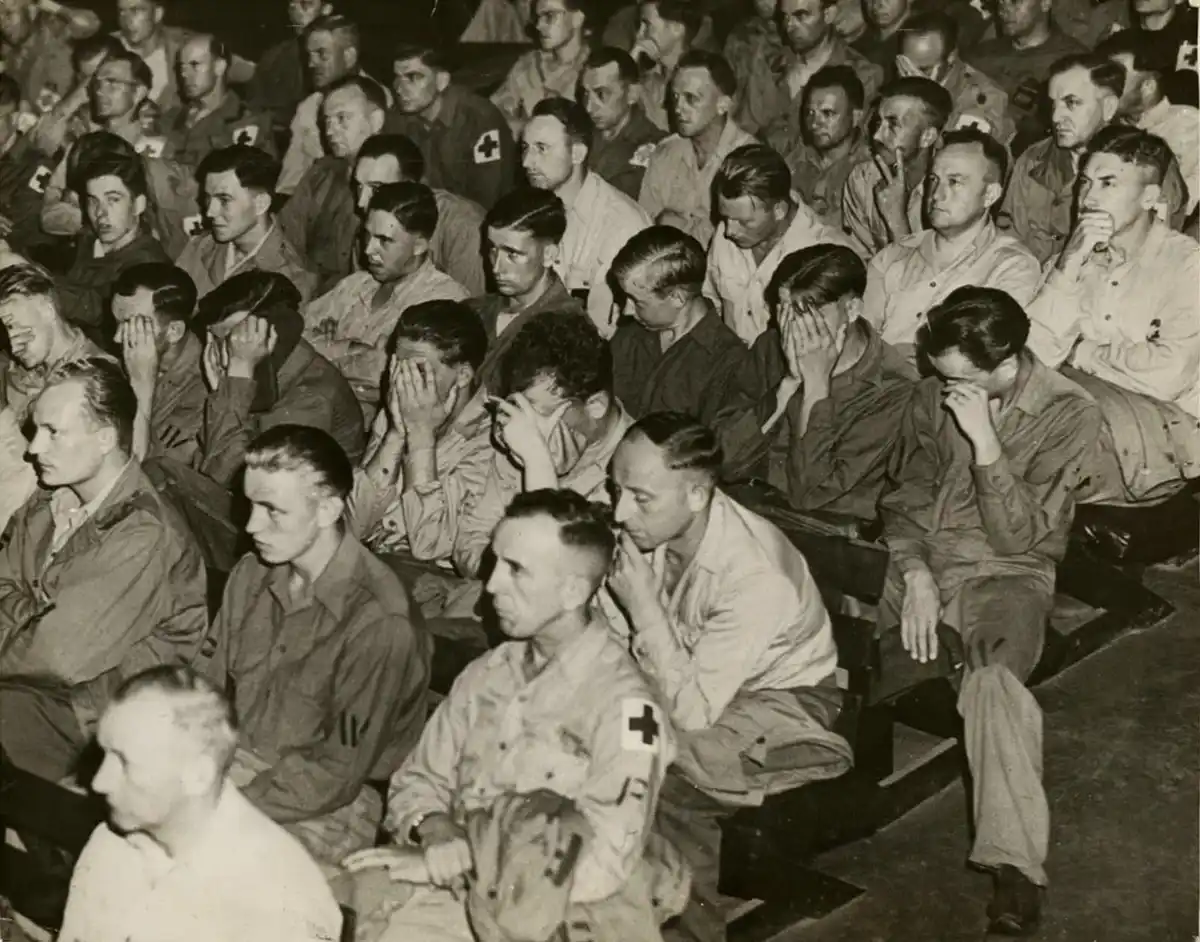 A black-and-white historical photo shows a crowd of men in military uniforms seated in rows, many covering their faces with…