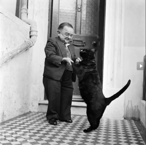 A black-and-white photo shows a man in a suit and glasses standing on a checkered floor, gently holding the paws of a black…
