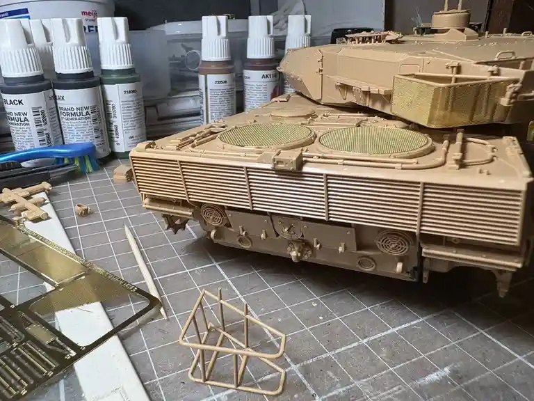 A partially assembled tan plastic tank model sits on a gridded work mat, surrounded by paint bottles, brushes, and…