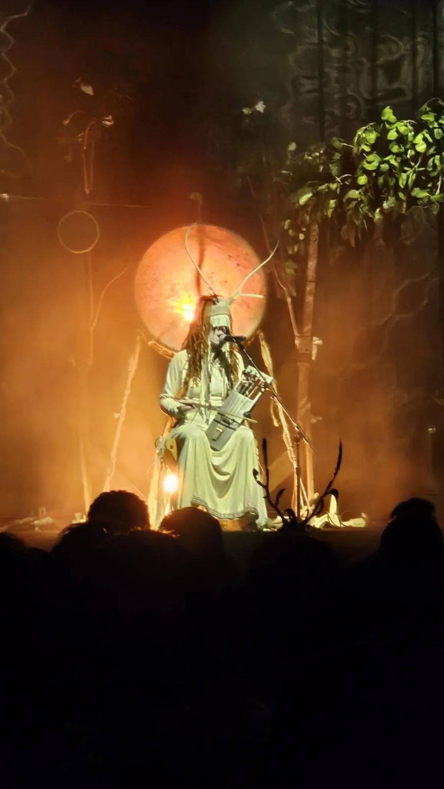 A person in a white robe and deer antler headpiece sits on a throne-like stage under warm orange lighting, holding a bow and…