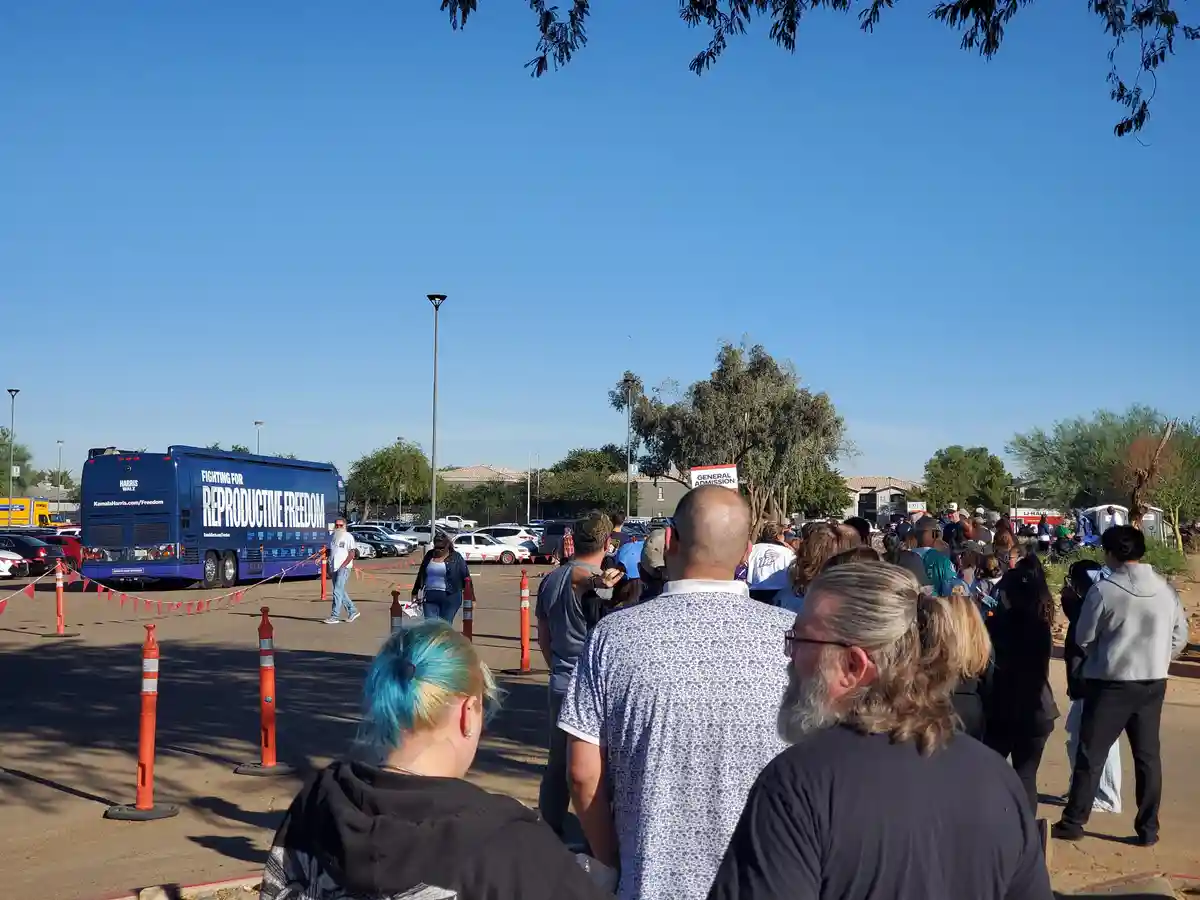 A crowd of people stands in a parking lot under a clear blue sky, facing a blue campaign bus with the text 'FIGHTING FOR…
