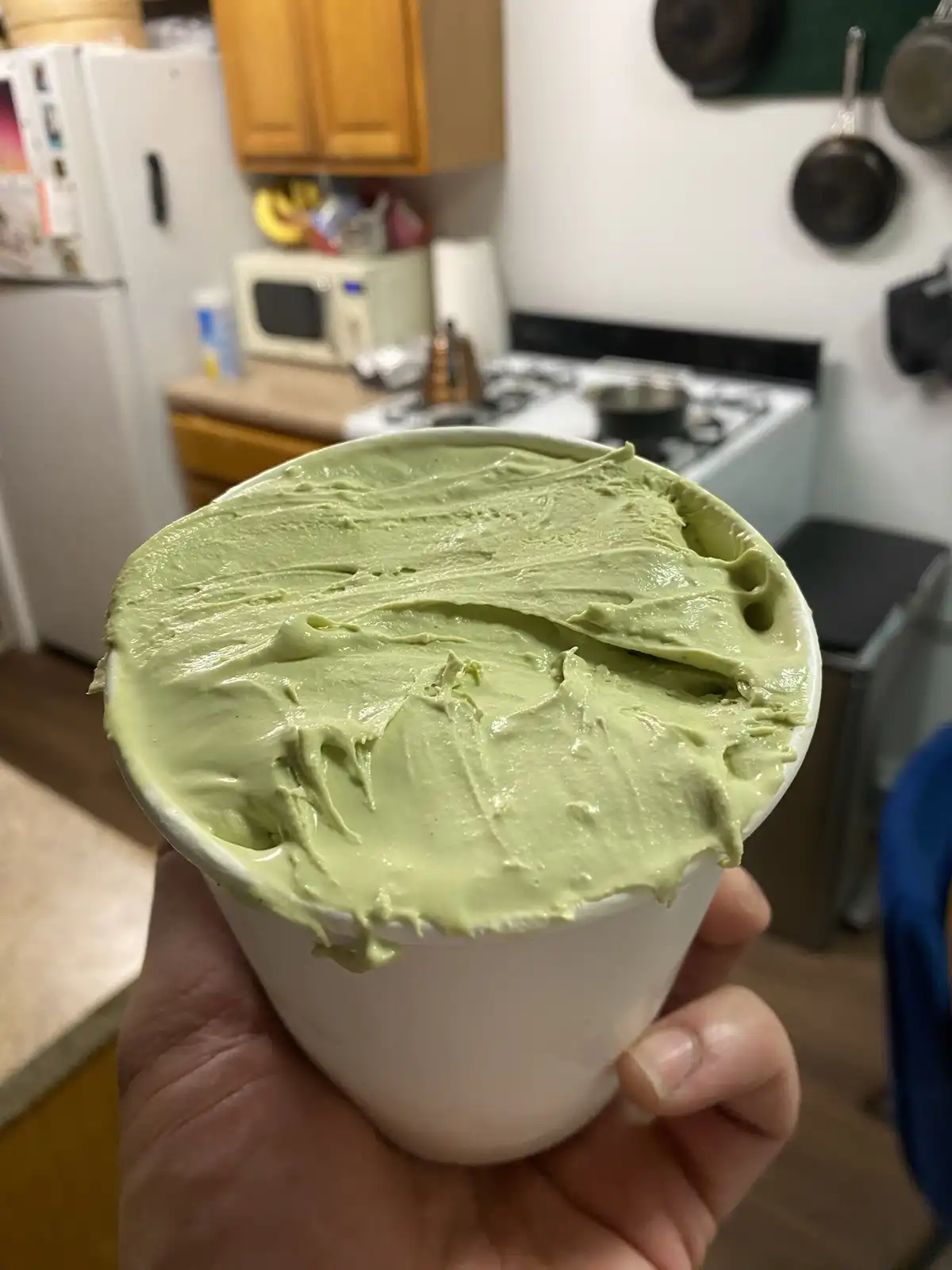 A hand holds a white cup overflowing with thick, light green matcha ice cream that has been scooped until it spills over the…