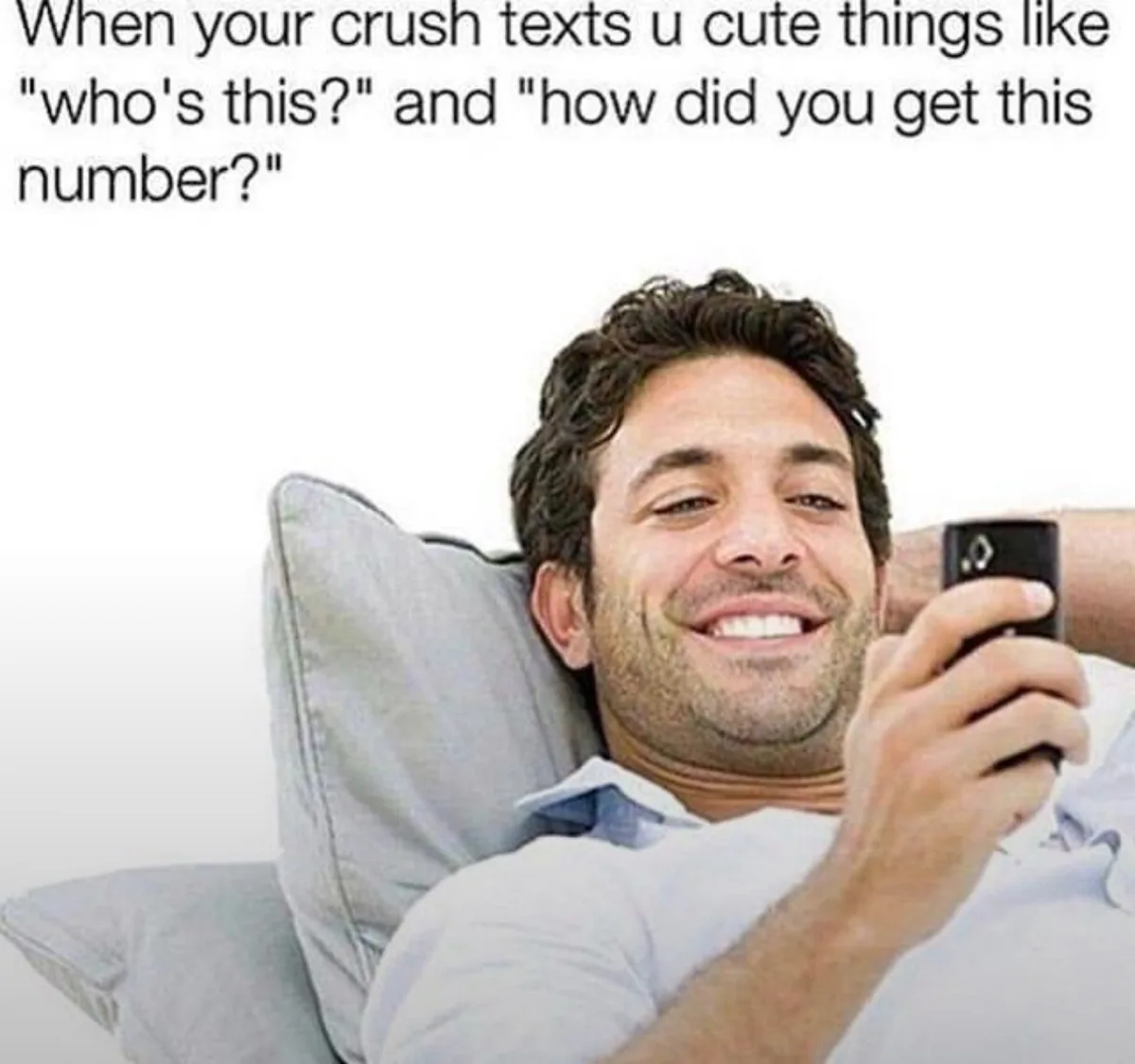 A man with dark hair and a light beard is lying back on a gray pillow, smiling broadly while holding a black smartphone in…
