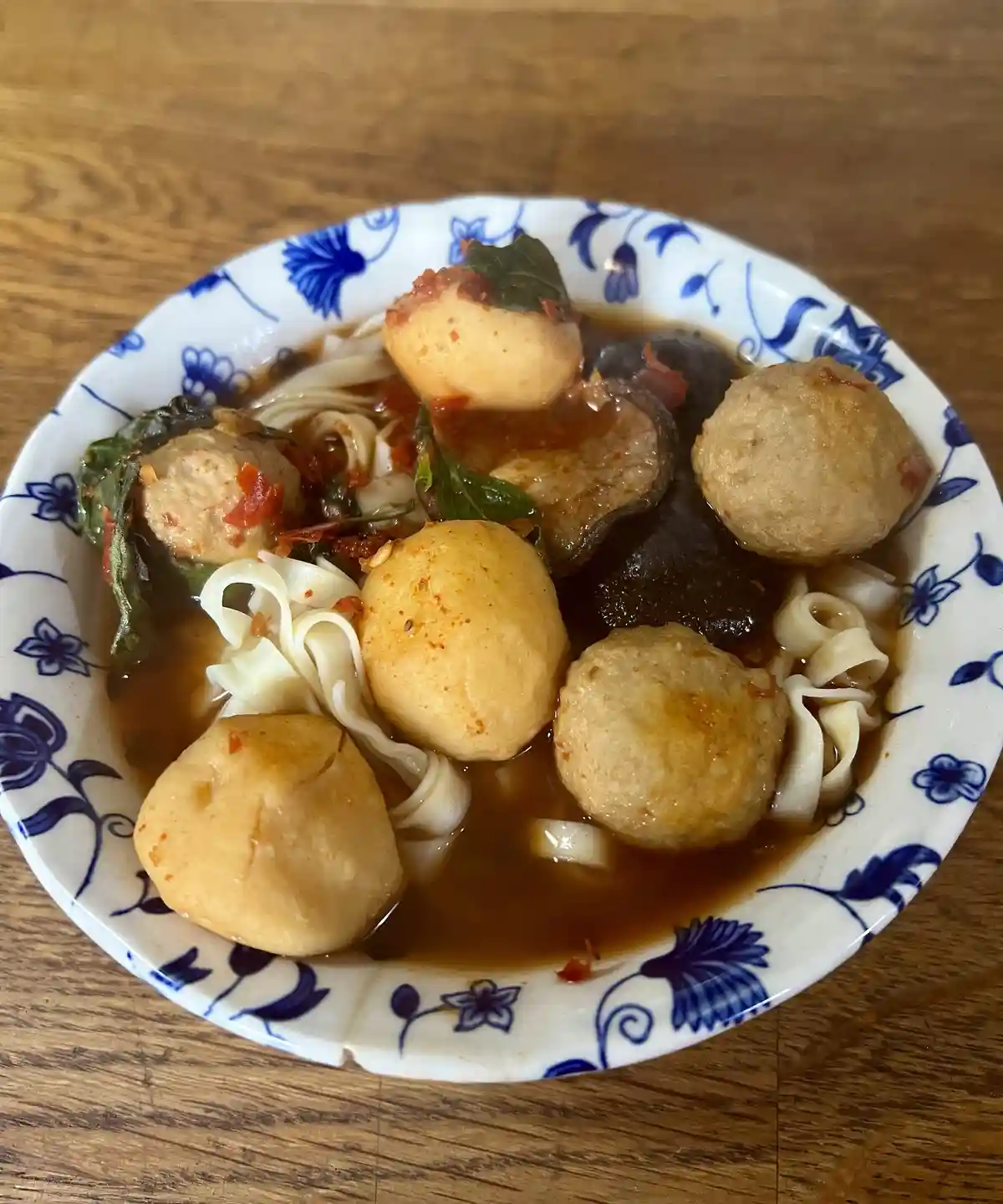 A close-up photo shows a bowl of noodle soup with several large, round, light-brown meatballs, dark mushrooms, green leaves…