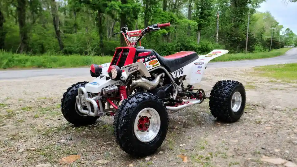 A red and white modified quad bike with large knobby tires and racing decals is parked on a gravel shoulder next to a paved…