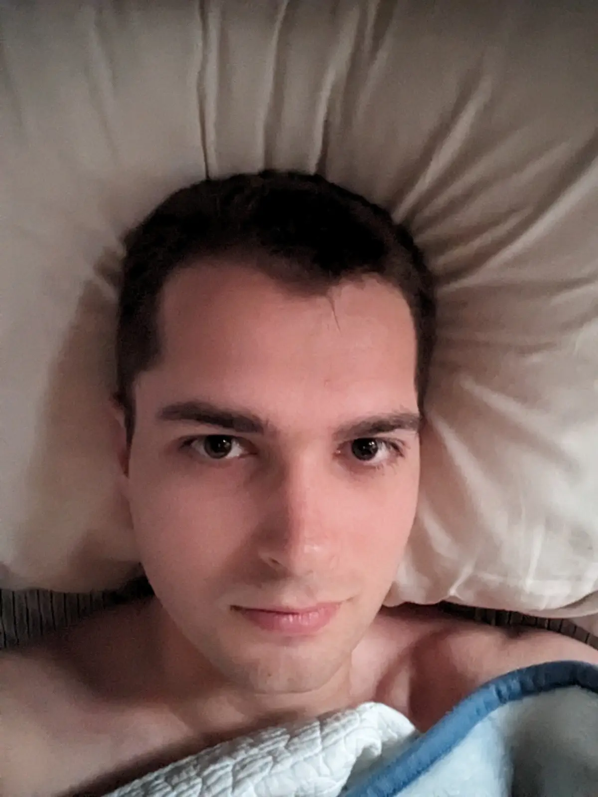 A young man with dark hair lies in bed, propped up on a white pillow, looking directly at the camera with a neutral…