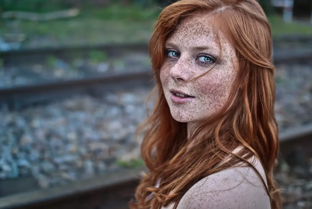A young woman with red hair and numerous freckles across her face and shoulders stands near train tracks. She looks directly…