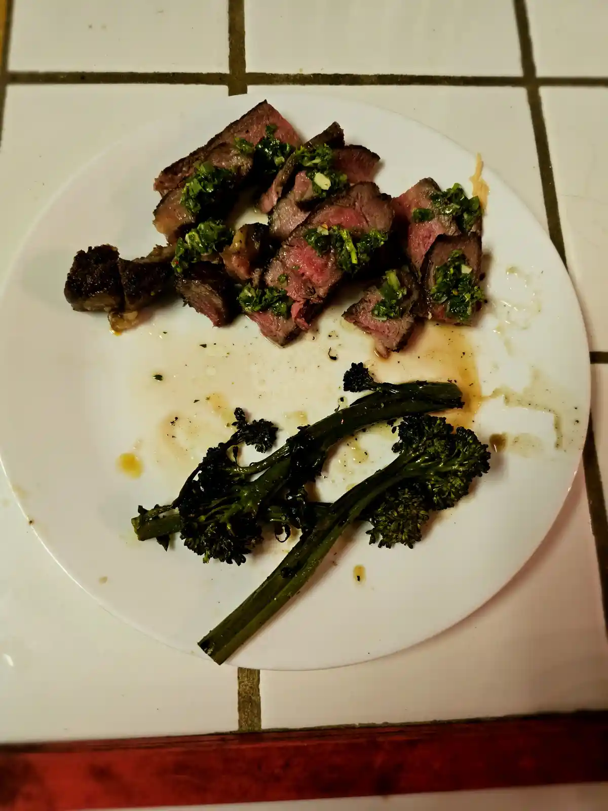 A white plate holds sliced medium-rare steak topped with green herb sauce and a few charred broccoli rabe stems, with most…