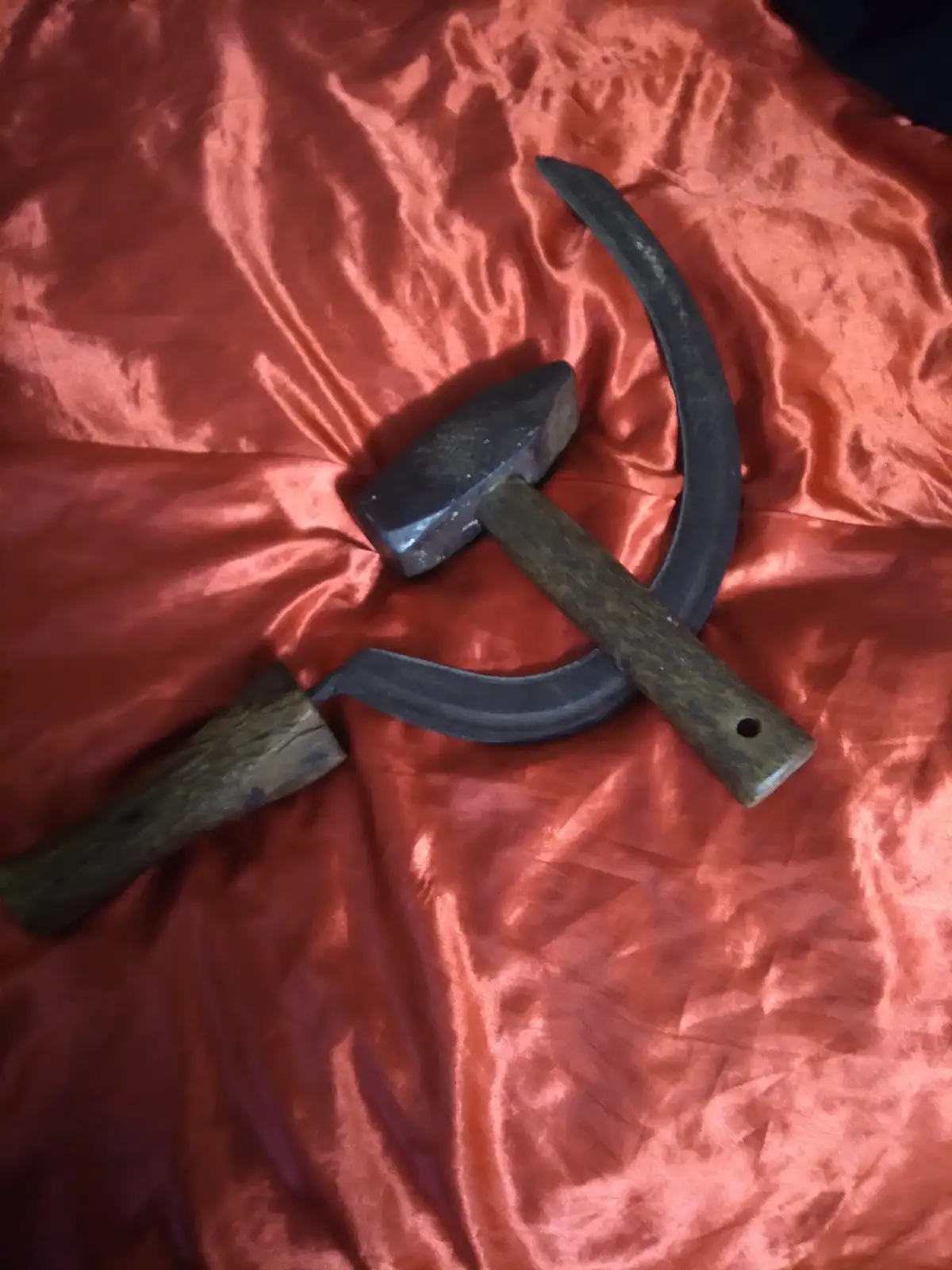 A hammer and a sickle, both with wooden handles, are crossed on a red satin fabric, forming the communist symbol. The…
